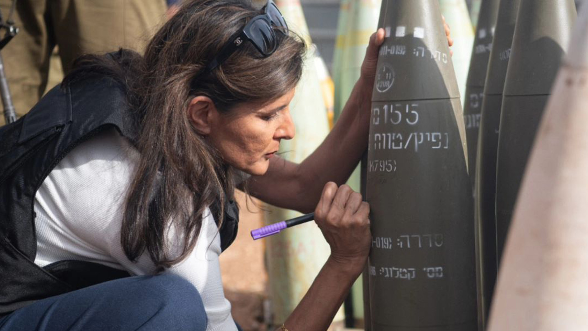 Side shot of Nikki Haley squatting down in a
    vest signing an artillery shell